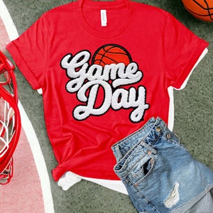 Puede incluir: Camiseta roja con un gráfico blanco de "Game Day" y un gráfico de baloncesto. La camiseta está sobre una superficie verde con un balón de baloncesto y un aro de baloncesto.