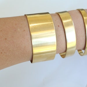 May include: Three gold-colored metal bracelets of varying widths are displayed on an arm. The bracelets are smooth and shiny, reflecting light. The widest bracelet is approximately 2 inches wide, while the narrowest is about 0.5 inches.