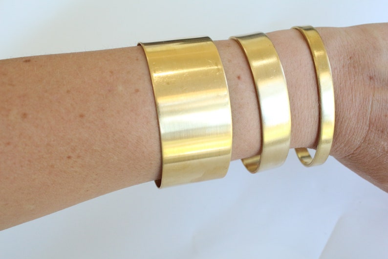 May include: Three gold-colored metal bracelets of varying widths are displayed on an arm. The bracelets are smooth and shiny, reflecting light. The widest bracelet is approximately 2 inches wide, while the narrowest is about 0.5 inches.