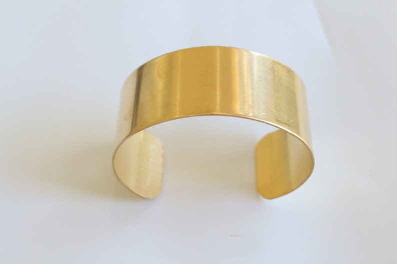 May include: A wide, gold-colored cuff bracelet. The open-ended bracelet has a smooth, polished surface and a simple, minimalist design. The bracelet is made of a shiny, metallic material.