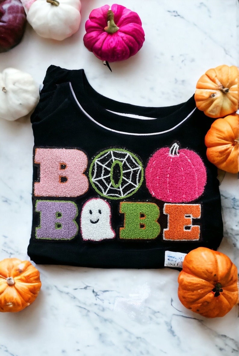 May include: Black long-sleeved shirt with a white trim around the neckline. The shirt has a festive Halloween design with the words "BOO BABE" in colorful, embroidered letters. The design also includes a spiderweb and a ghost.