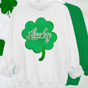 Könnte beinhalten: Weißer Pullover mit einem grünen vierblättrigen Kleeblatt-Design und dem Wort "Lucky" in einer glitzernden Schrift.