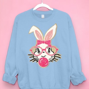 Könnte beinhalten: Ein hellblauer Pullover mit einem weißen Hasen, der eine rosa Brille und eine rosa Schleife trägt und eine rosa Blase bläst. Der Hase ist aus Glitzer.