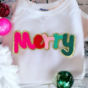 May include: White t-shirt with a colorful "Merry" embroidered patch. The letters are pink, red, light pink, green, and dark green with gold outlining.
