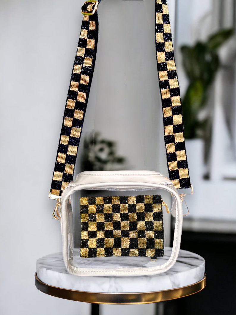 May include: A clear plastic crossbody bag with a white trim and a black and gold checkered beaded strap. The bag has a zipper closure and a clear compartment that shows a black and gold checkered beaded pouch.