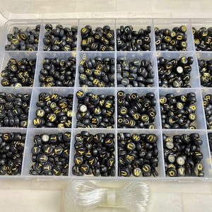 May include: A clear plastic storage box filled with black letter beads with gold lettering. The beads are organized by letter in individual compartments. A clear elastic cord is included.