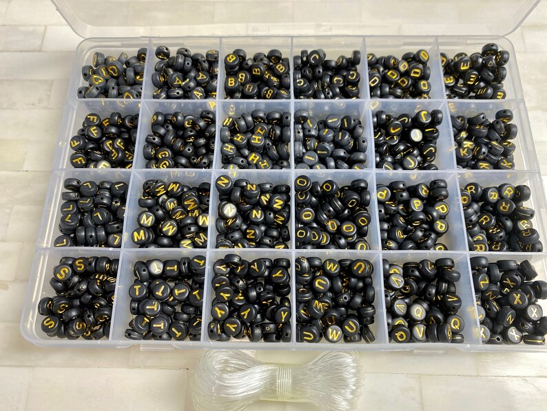 May include: A clear plastic storage box filled with black letter beads with gold lettering. The beads are organized by letter in individual compartments. A clear elastic cord is included.