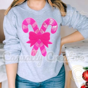 May include: A gray sweatshirt with a pink candy cane and bow design. The design is made of sequins and is a festive Christmas design.