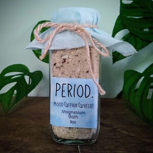 Könnte beinhalten: Ein klares Glas mit Badesalz, beschriftet mit "PERIOD. MOOD PAIN RELIEF Magnesium Bath 9oz." Das Glas ist mit einem hellblauen Stoff verschlossen und mit Bindfaden gebunden. Der Hintergrund zeigt grüne Blätter.