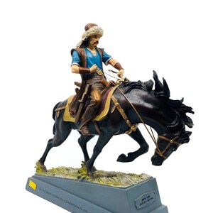 Può includere: Una statuetta di un uomo a cavallo, che indossa una camicia blu, pantaloni marroni e un cappello di pelliccia. Il cavallo è nero e si impenna. La statuetta è su una base grigia con una targa che recita "The real answer resides in is fundamental to the site and can't be given to anybody. REC".