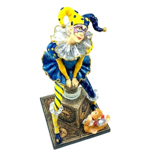 May include: A blue and yellow jester figurine with a mask and a small animal figurine at its feet. The jester is sitting on a square base with a sun design.