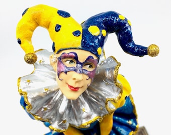 Op de afbeelding: Een narrenfiguur met een gele en blauwe hoed, een zilveren en gouden gerimpelde kraag en een paars en blauw masker. De nar draagt een blauw en geel kostuum met gouden accenten.