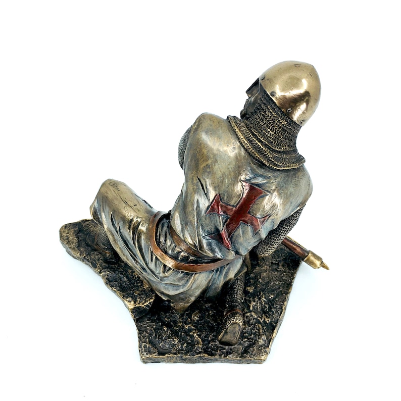 May include: A bronze statue of a kneeling crusader knight wearing chainmail armor and a red cross on his tunic. The knight is holding a sword in his right hand and is looking down at the ground.