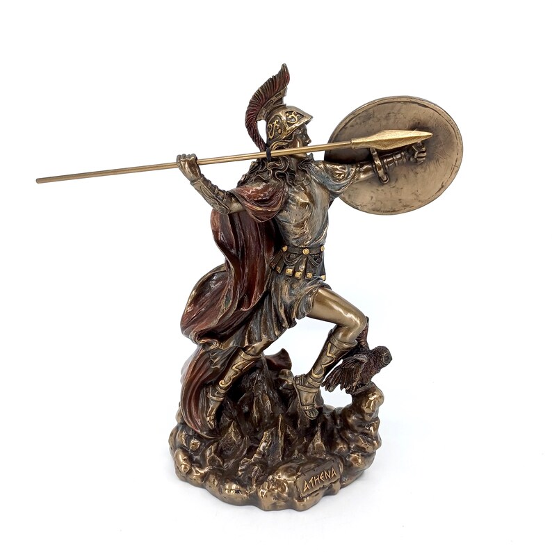 Puede incluir: Una estatua de bronce de Atenea, la diosa griega de la sabidur&iacute;a y la guerra, sosteniendo una lanza y un escudo. Est&aacute; de pie sobre una base rocosa con un b&uacute;ho a sus pies. La estatua est&aacute; pintada en tonos de marr&oacute;n, dorado y rojo.