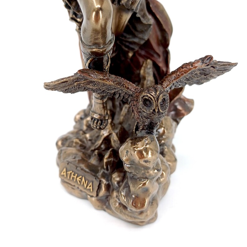 Puede incluir: Estatua de bronce de la diosa griega Atenea, de pie sobre una base rocosa con un b&uacute;ho posado en su mano. La estatua est&aacute; inscrita con la palabra "ATHENA" en la base.