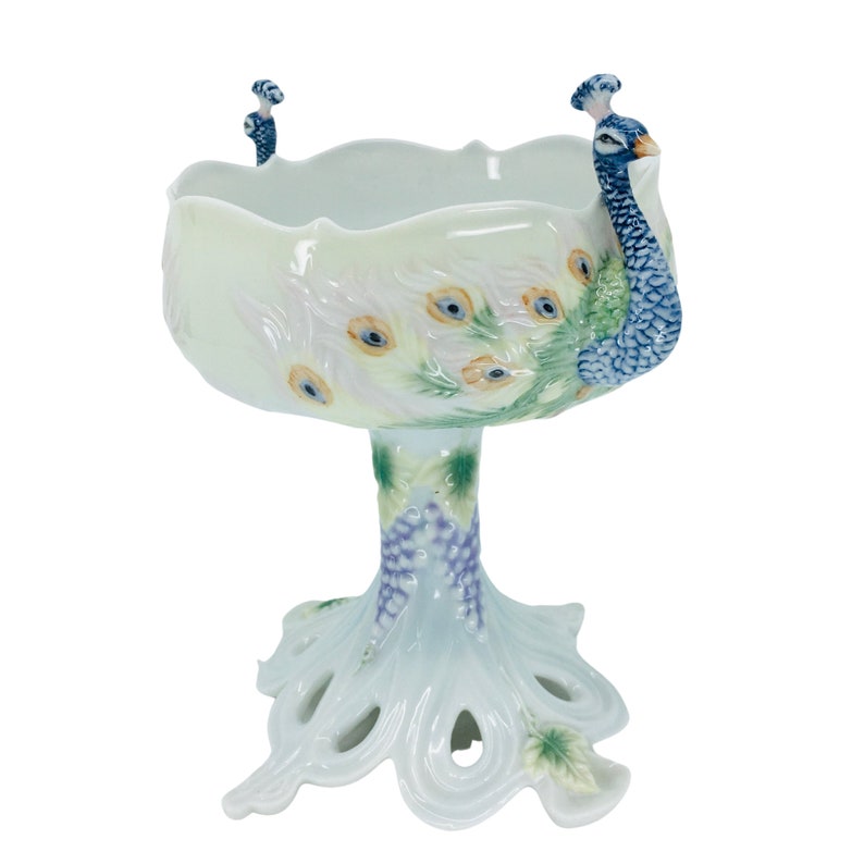 May include: A decorative, off-white ceramic peacock bowl. The bowl features a scalloped rim and is supported by a pedestal base. The peacock's feathers are painted with blue, green, and yellow accents. The bowl is approximately 10 inches tall.