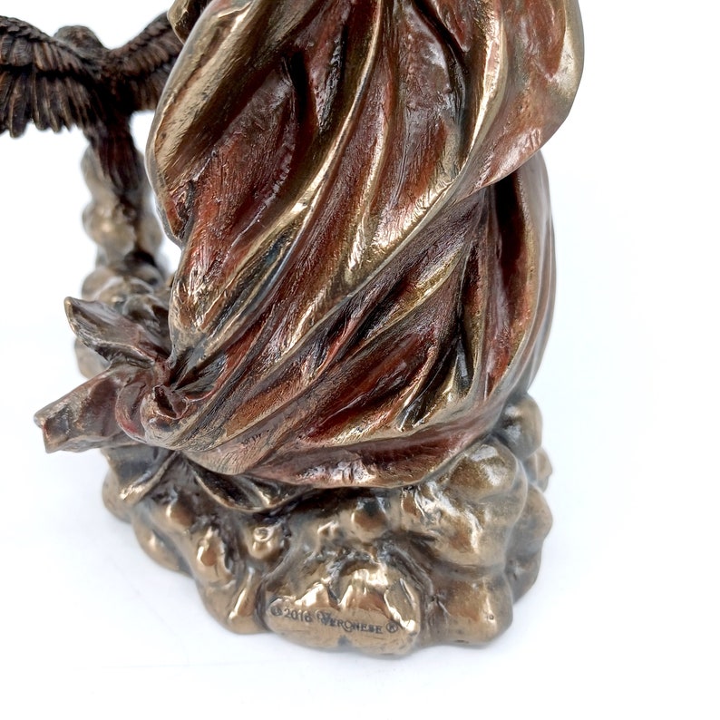 Puede incluir: Una escultura de bronce de una mujer con ropas fluidas y un &aacute;guila posada en su brazo. La escultura est&aacute; colocada sobre una base rocosa y tiene una inscripci&oacute;n de copyright en la parte inferior.
