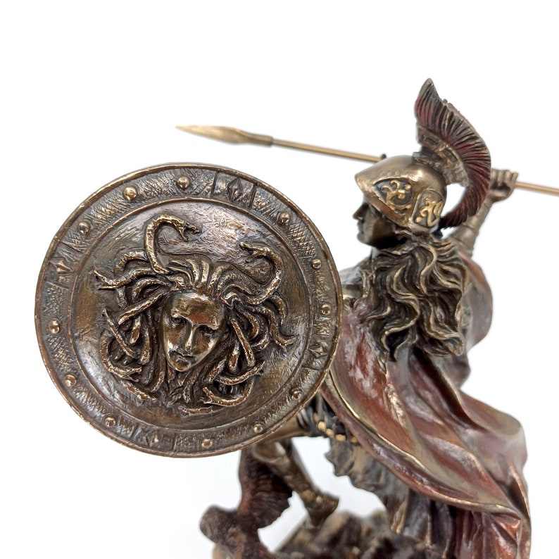 Puede incluir: Una estatua de bronce de un guerrero griego, sosteniendo una lanza y un escudo con la cabeza de Medusa en &eacute;l. El guerrero lleva un casco y una capa roja.