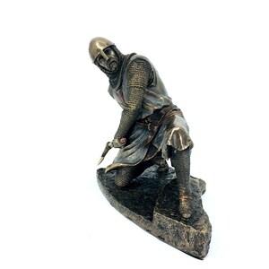 May include: A bronze statue of a kneeling knight in chainmail armor, holding a sword. The knight is wearing a helmet and has a red cross on his chest. The statue is on a rocky base.