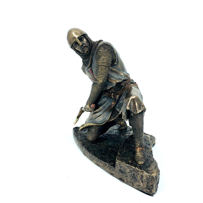 May include: A bronze statue of a kneeling knight in chainmail armor, holding a sword. The knight is wearing a helmet and has a red cross on his chest. The statue is on a rocky base.