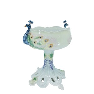 May include: A white ceramic peacock pedestal bowl. The bowl features two peacock figures on the rim and a detailed peacock feather design. The base is shaped like a peacock's foot, with a grape vine design on the stem.