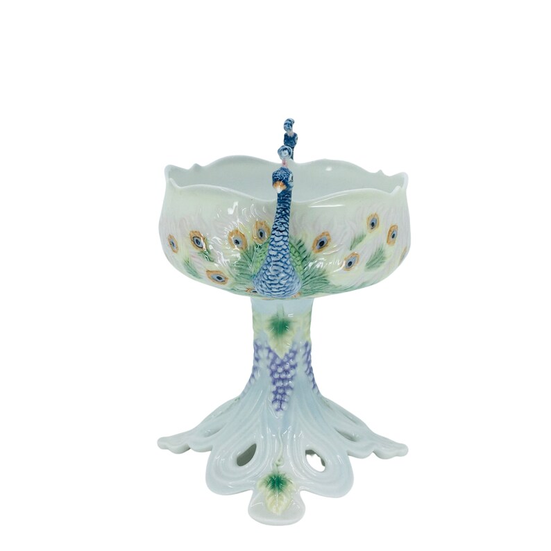 May include: A white ceramic pedestal bowl with a peacock design. The bowl features a scalloped rim and is decorated with peacock feather motifs in yellow, blue, and green. A blue peacock figure serves as a handle. The base is shaped like a peacock's tail.