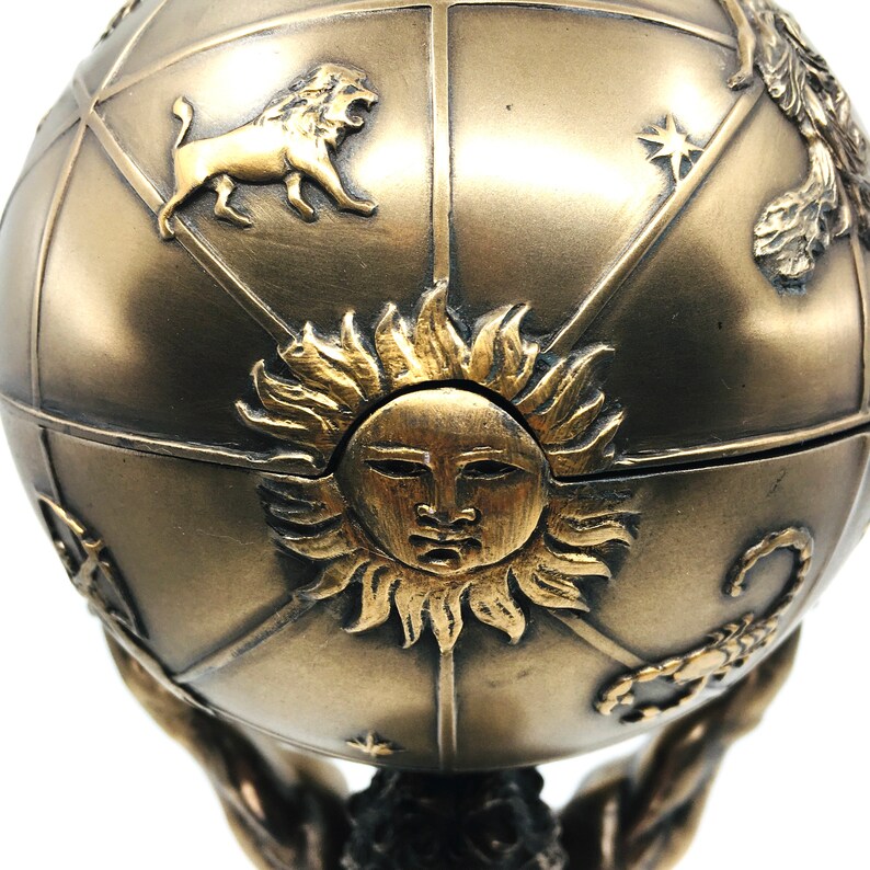 May include: A gold-toned metal globe with a sun face and zodiac symbols. The globe is held up by two hands.