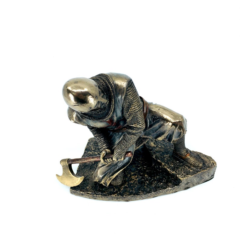 May include: A bronze figurine of a kneeling knight in chainmail armor holding an axe. The knight is wearing a red sash and has a helmet on his head. The figurine is set on a rocky base.