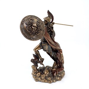 Puede incluir: Una estatua de bronce de una diosa griega sosteniendo una lanza y un escudo con la cabeza de Medusa en &eacute;l. La diosa est&aacute; de pie sobre una base rocosa con dos b&uacute;hos a sus pies.