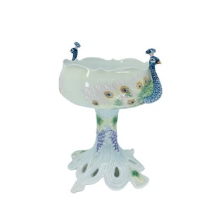 May include: A decorative ceramic peacock pedestal bowl. The bowl is white with blue, green, and yellow accents. The base is shaped like a peacock's tail. The bowl features two peacock heads on either side.