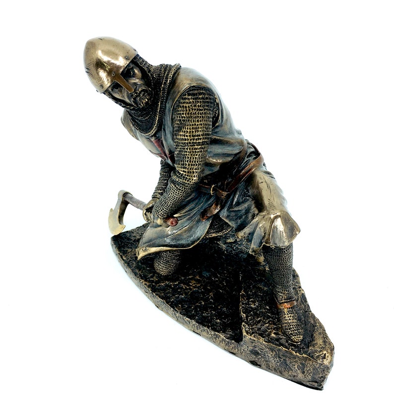 May include: A bronze statue of a kneeling knight in chainmail armor holding an axe. The knight is wearing a helmet with a red cross on the front. The statue is set on a rocky base.