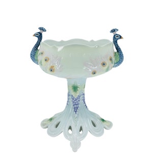 May include: A white ceramic peacock bowl with a scalloped rim. Two peacock figures are on the sides. The bowl has a pedestal base with a grape and leaf design. The base is shaped like a peacock's tail.