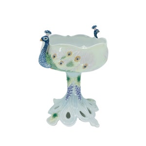 May include: A white ceramic peacock bowl with a pedestal base. The bowl features a peacock design with blue, green, and yellow accents. The base is shaped like a peacock's foot.