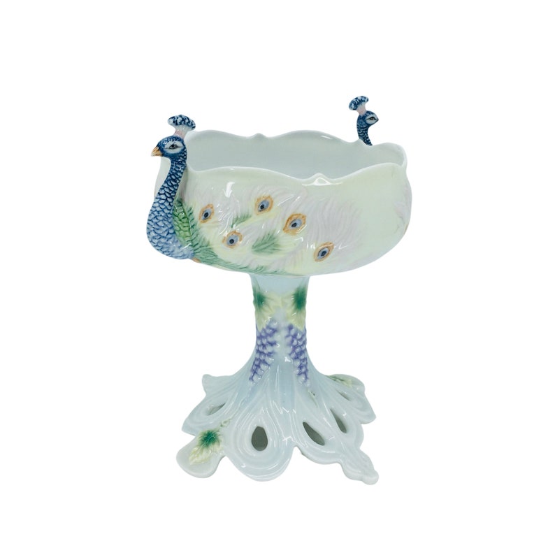 May include: A white ceramic peacock bowl with a pedestal base. The bowl features a peacock design with blue, green, and yellow accents. The base is shaped like a peacock's foot.