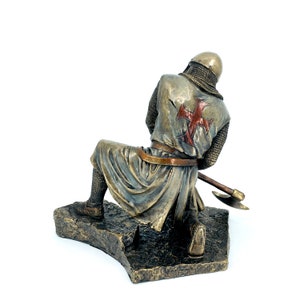 May include: A bronze statue of a kneeling knight wearing chainmail armor and a red cross on his chest. He is holding an axe in his right hand. The statue is on a rocky base.
