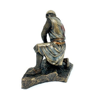 May include: A bronze statue of a kneeling crusader knight wearing chainmail armor and a red cross on his tunic. The statue is on a rocky base.