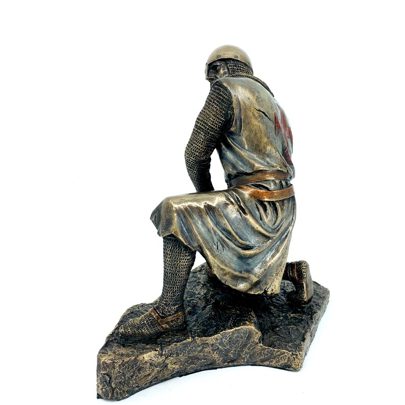 May include: A bronze statue of a kneeling crusader knight wearing chainmail armor and a red cross on his tunic. The statue is on a rocky base.