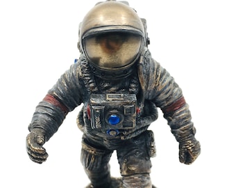 Op de afbeelding: Een bronzen astronautbeeldje met een blauw en rood geaccentueerd ruimtepak, staand op een getextureerde maanbodem.
