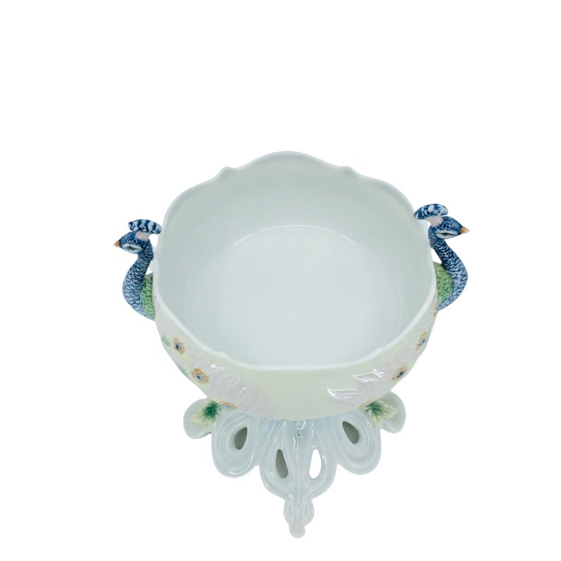 May include: A white ceramic bowl with a scalloped rim, featuring peacock handles and a decorative base. The handles are detailed with blue, green, and white peacock designs. The base has a peacock feather design.