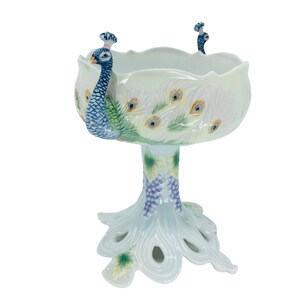 May include: A white ceramic peacock bowl with a scalloped rim. The bowl features a peacock design with blue, green, and yellow accents. The base is shaped like a peacock's tail, and the bowl is supported by a pedestal.