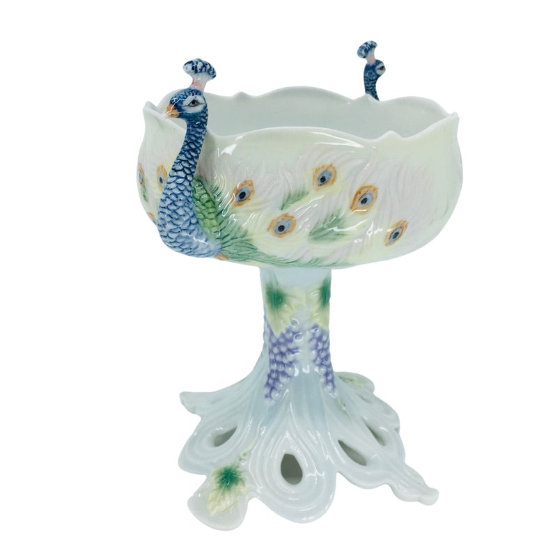 May include: A white ceramic peacock bowl with a scalloped rim. The bowl features a peacock design with blue, green, and yellow accents. The base is shaped like a peacock's tail, and the bowl is supported by a pedestal.