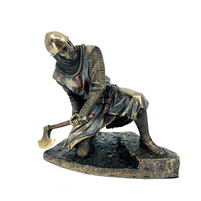 May include: A bronze statue of a kneeling crusader knight holding an axe. The knight is wearing chainmail armor and a red cross on his chest. The statue is on a base that looks like a rock.