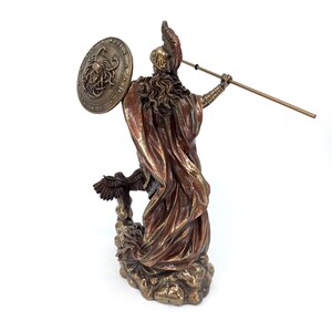 Puede incluir: Una estatua de bronce de una diosa griega, posiblemente Atenea, de pie sobre una base en forma de nube. Ella sostiene una lanza en su mano derecha y un escudo con una cabeza de Medusa en su mano izquierda. La estatua est&aacute; pintada en tonos dorados y marrones.