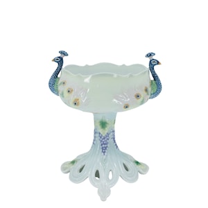 May include: A white ceramic pedestal bowl with a scalloped rim, featuring two peacock figures on the sides. The bowl's base is adorned with a grape vine design and peacock feather accents, creating an elegant decorative piece.