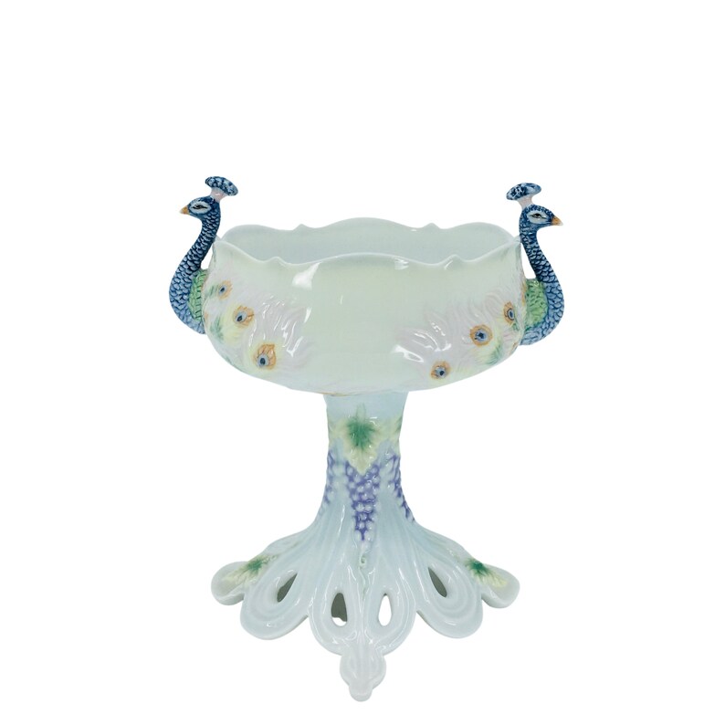 May include: A white ceramic pedestal bowl with a scalloped rim, featuring two peacock figures on the sides. The bowl's base is adorned with a grape vine design and peacock feather accents, creating an elegant decorative piece.