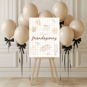 May include: A sign on a wooden easel with the words "Welcome to Friendsgiving" in brown script. The sign is decorated with line drawings of food and fall elements. Beige balloons with black bows are arranged around the sign.