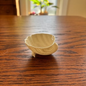 May include: A small, off-white ceramic dish shaped like a seashell, featuring a scalloped edge and a smooth, glossy finish. The bowl is supported by three small, delicate feet, giving it a raised appearance on the surface it rests upon.