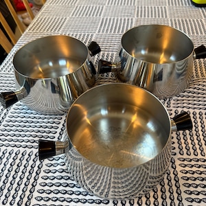 Op de afbeelding: Drie roestvrijstalen kookpotten met zwarte handvatten staan op een tafelkleed met patroon. De potten hebben een glanzend, reflecterend oppervlak en een afgeronde vorm. De handvatten zijn cilindrisch en aan de zijkanten van de potten bevestigd.