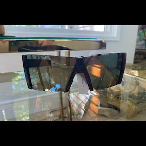 Peut inclure: Une paire de lunettes de soleil brun foncé avec une monture carrée et des verres foncés. Les lunettes de soleil sont posées sur une surface en verre. L'étiquette sur les lunettes de soleil indique "Fabriqué en Italie".
