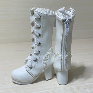 Puede incluir: Un par de botas blancas de muñeca con cordones en la parte delantera y cierre de cremallera en la parte trasera. Las botas tienen un tacón pequeño y ribete decorativo con volantes. Las botas miden aproximadamente 10 cm de alto.